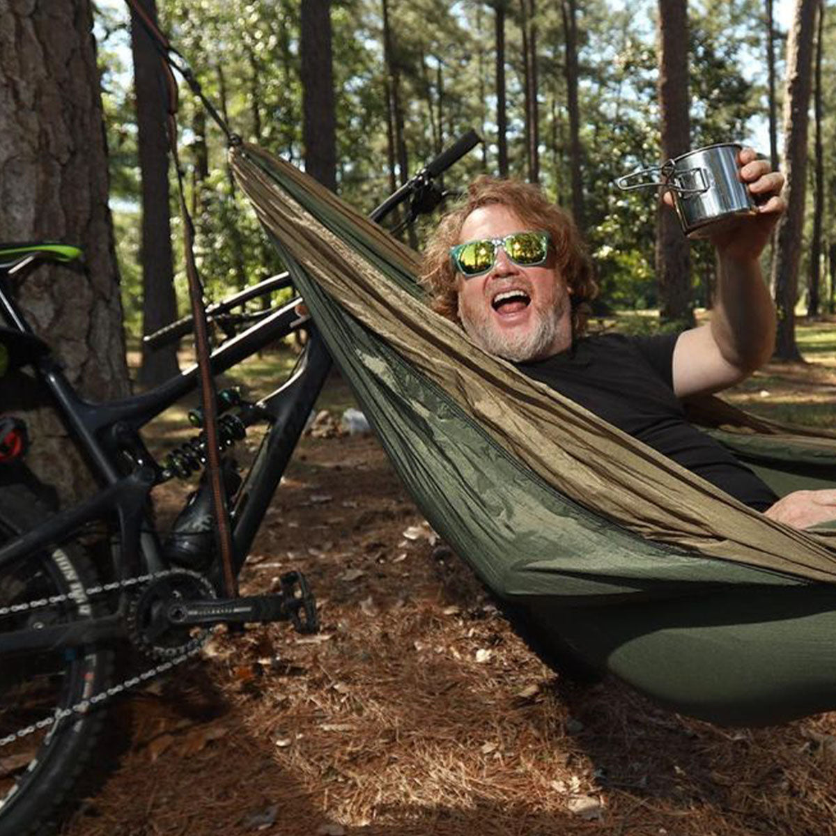 man in hammock wearing sunglasses holding mug in front of mountain bike