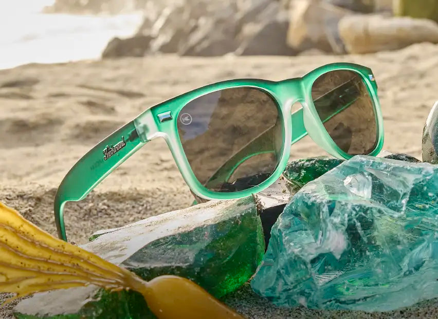 Glass Beach Fort Knocks sunglasses photographed on a sandy beach among green glass rocks and seashells.
