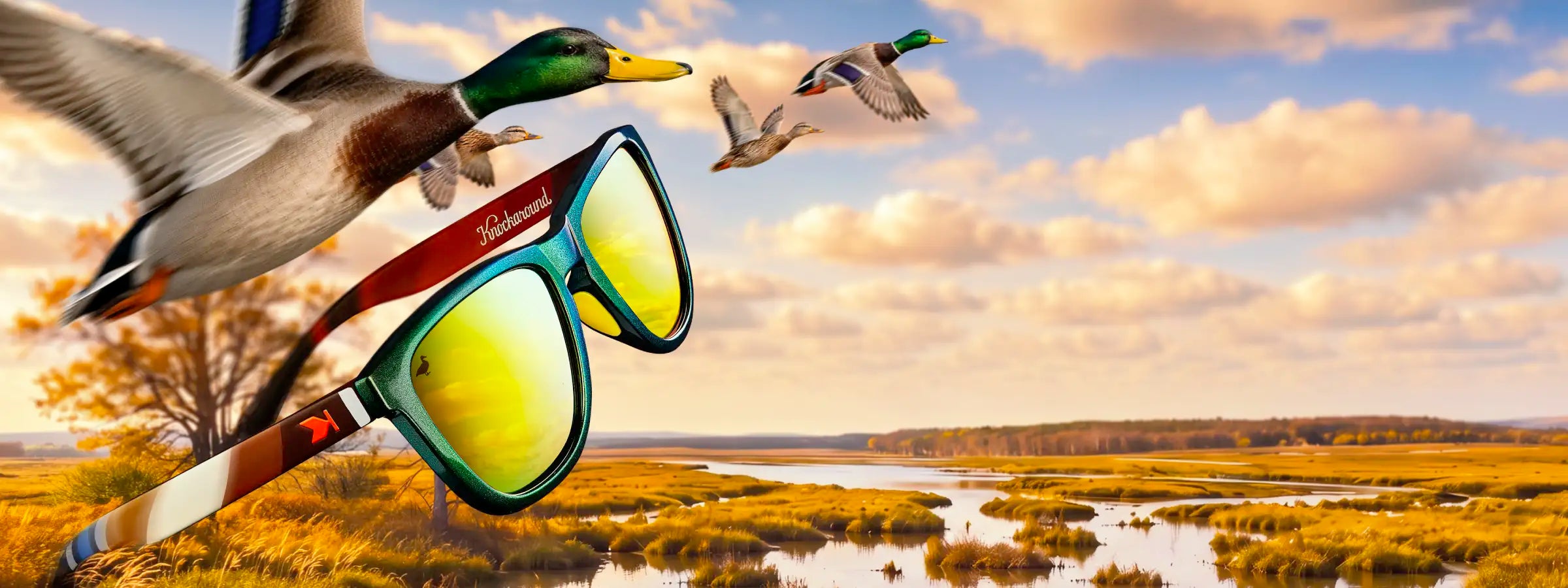 A pair of Mallard Limited Edition Premiums Sport sunglasses with mirrored yellow lenses and a mallard duck pattern on the frame is shown flying over a marshland landscape with flying mallards in the background.