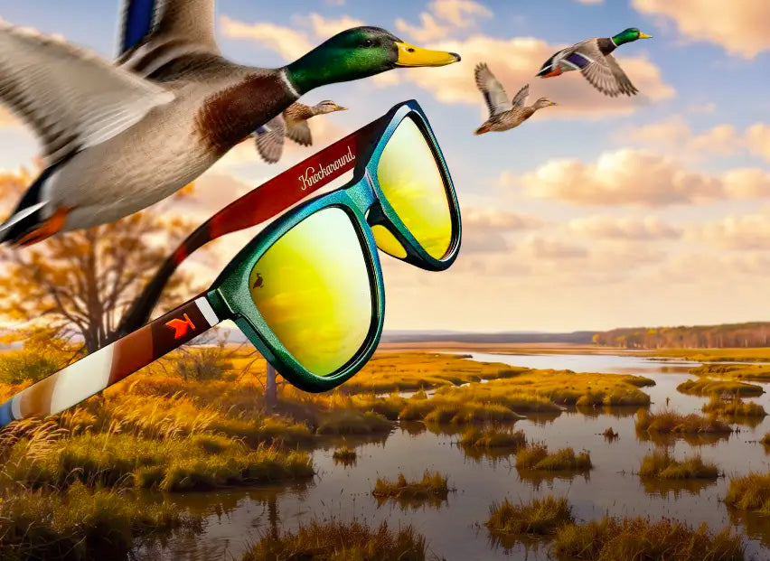 A pair of Mallard Limited Edition Premiums Sport sunglasses with mirrored yellow lenses and a mallard duck pattern on the frame is shown flying over a marshland landscape with flying mallards in the background.