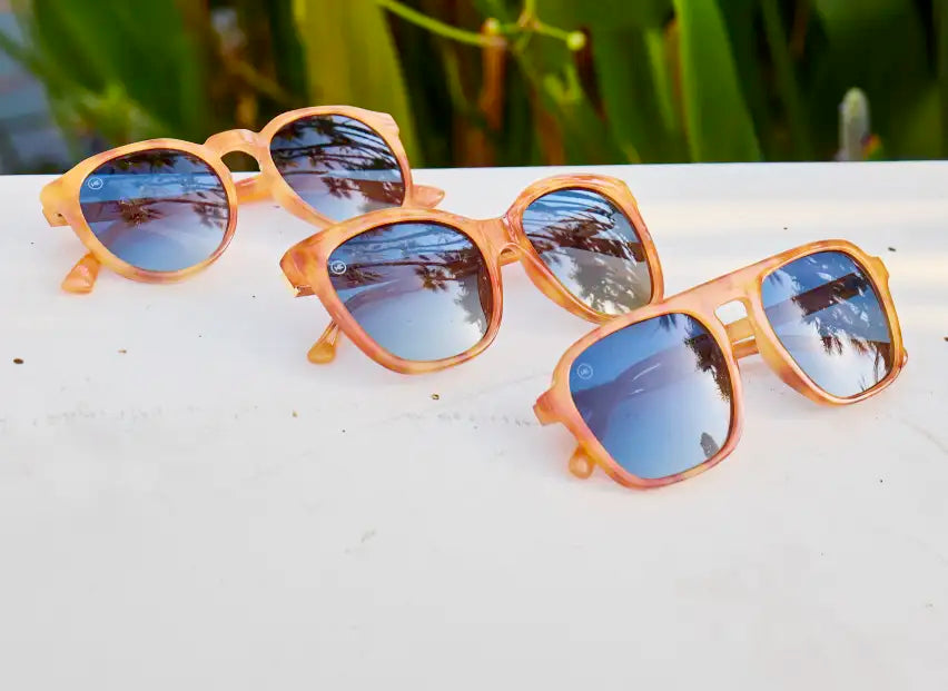 3 pairs of Sienna Tortoise Shell Knockaround sunglasses. Tortoise shell frame with blue lenses photographed on a granite surface with plants in the background