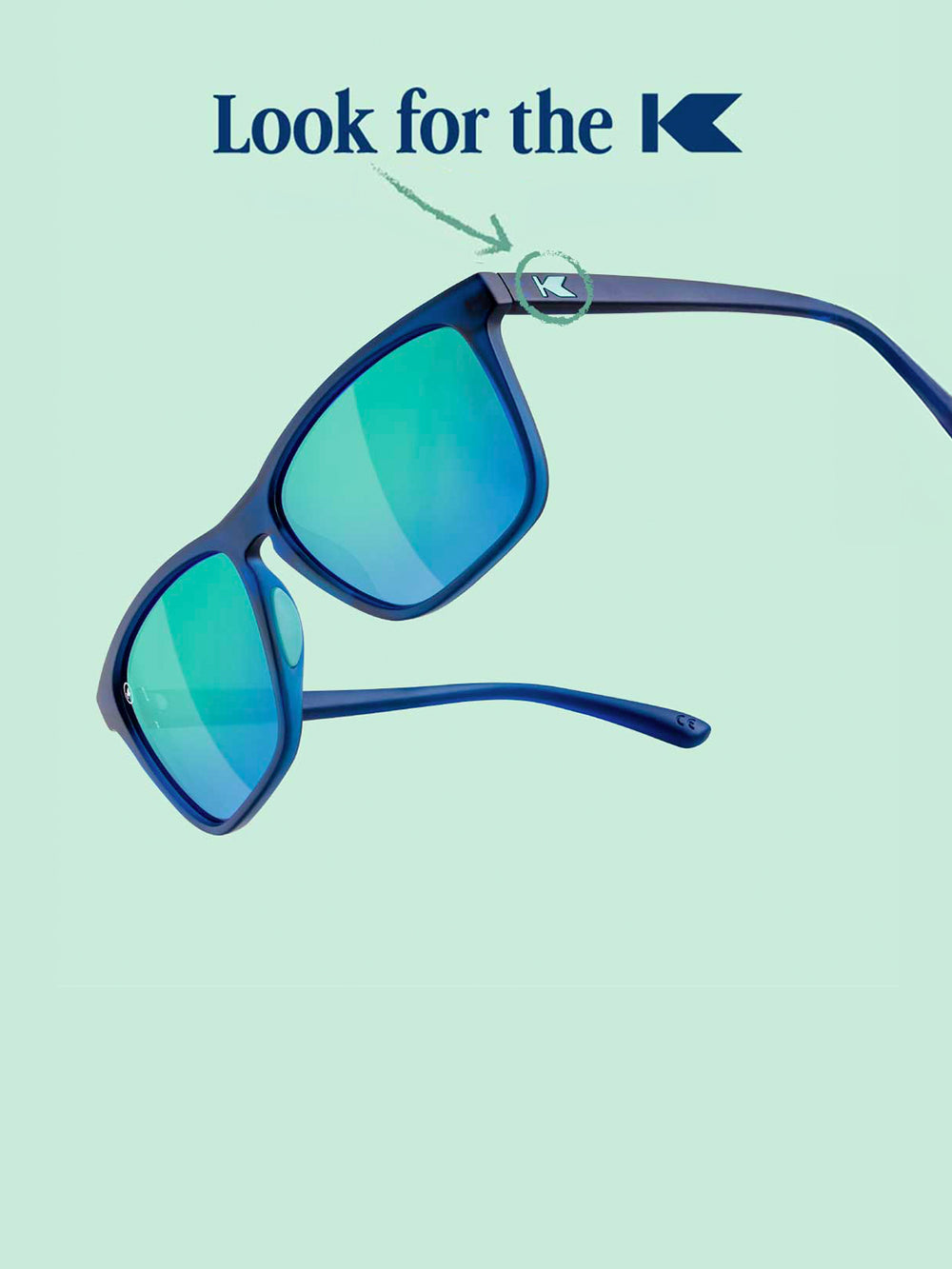 Navy / Mint Fast Lanes Sport sunglasses photographed on a light blue background with "Look for the K" text overlayed and an arrow pointing to the K logo on the sunglasses