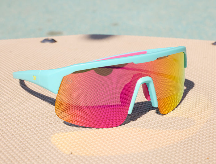 A pair of Volts wraparound sunglasses with a light blue frame and a vibrant pink-to-yellow mirrored lens, resting on a textured beige surface.