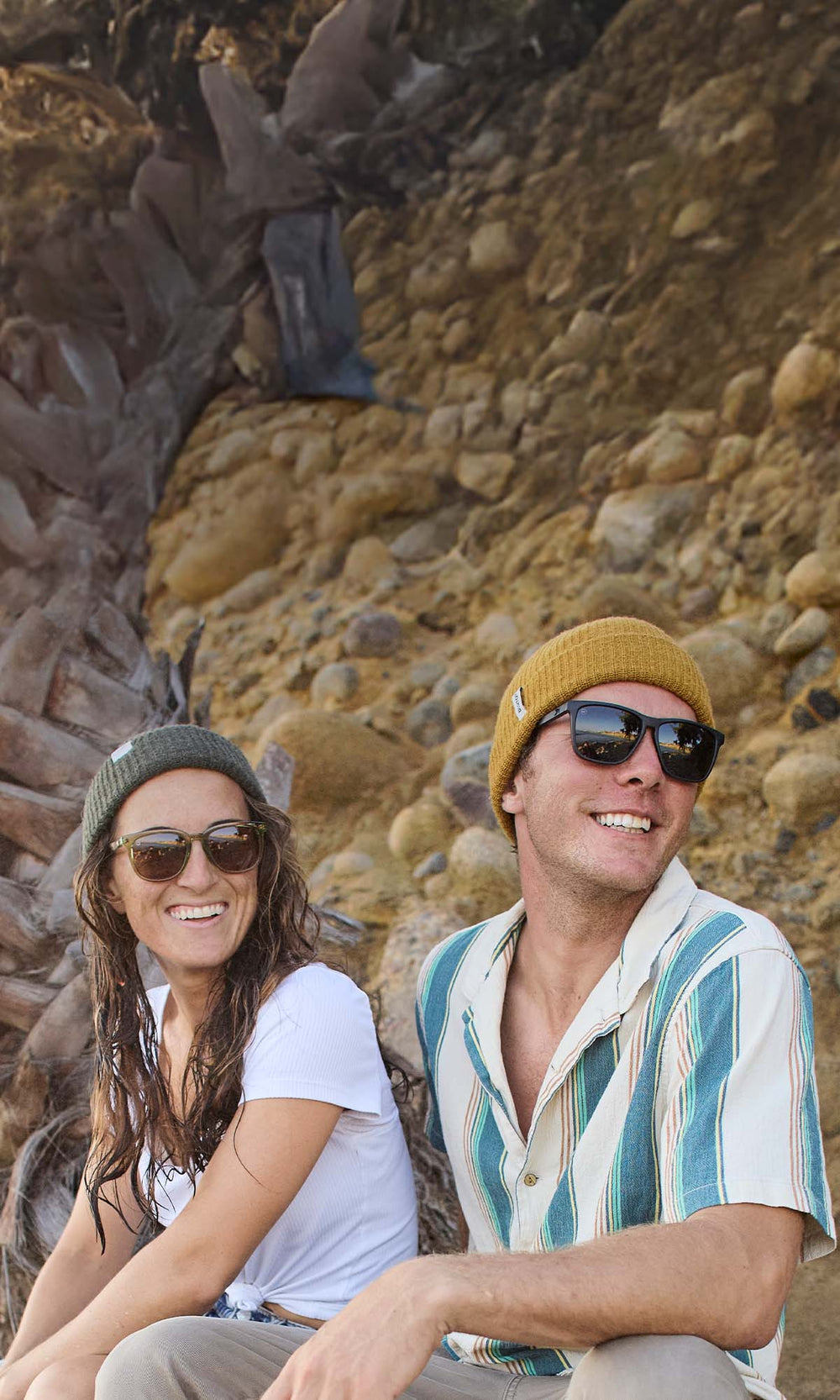Two people sitting on a rocky beach wearing Knockaround sunglasses.